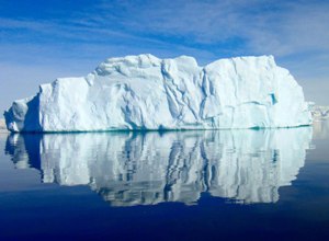 A large iceberg floating in a calm blue ocean, reflecting symmetrically on the water's surface under a clear sky.