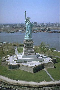 Aerial view of the Statue of Liberty standing on a star-shaped base surrounded by green fields with the cityscape and river in the background.
