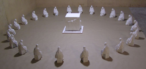 An artistic installation featuring twenty small draped figurines symmetrically arranged in a circle around a central illuminated square on the floor, in a room with subdued lighting.