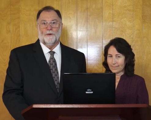 A man and a woman standing behind a podium with a laptop. The man is wearing a dark suit and tie, and the woman is in a purple blouse. They are positioned in front of a wooden panel wall.