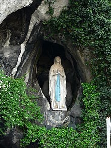 A statue of the Virgin Mary standing serenely in a natural cave niche, surrounded by lush green foliage. The figure is depicted with hands joined in prayer, wearing a long robe, with a sash and a halo. Below the statue, a symbol representing the constellation Virgo is visible, subtly integrated into a design platform that supports the statue.