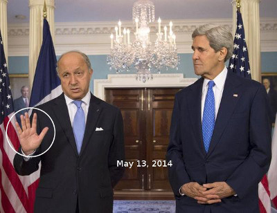 Two men in suits standing in a room adorned with flags and a chandelier, engaging in a conversation on May 13, 2014. The man on the left is gesturing with an open hand.
