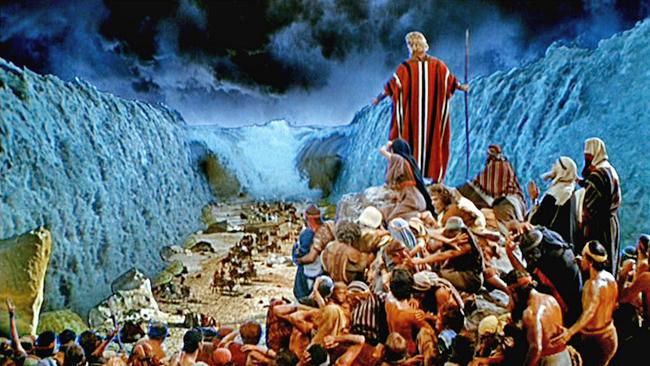 A cinematic depiction of a biblical scene where a leader in a striped red and white robe stands elevated on a rocky path, parting a massive blue sea with a staff. A large group of people in ancient attire gather around him, some looking back towards the towering walls of water on either side under a stormy sky.