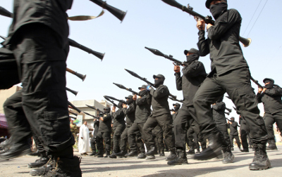 A group of individuals in black uniforms and boots marching in a parade, each carrying a rifle held vertically in front of them.