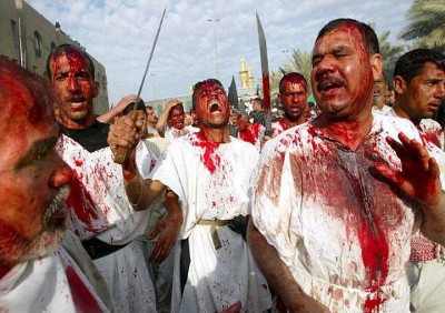 A group of men involved in a religious procession, covered in red paint to symbolize blood, some holding bladed weapons, expressing intense emotion under a clear sky.