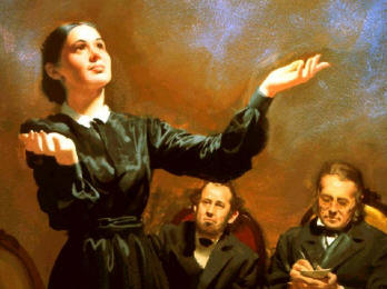 A woman in a black dress stands with her arms raised, gesturing skyward, as she elegantly explains the Mazzaroth. Two men sit behind her, observing attentively; one holds a small book and the other looks pensive.