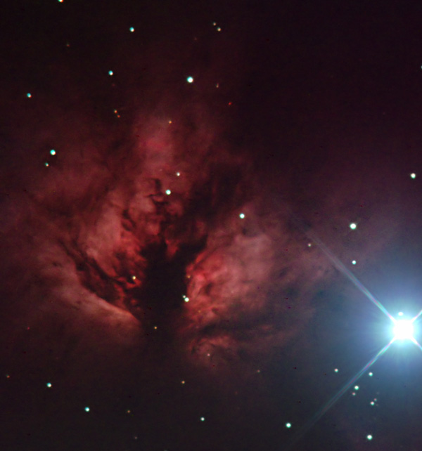 A brilliant celestial display shows a large, luminous star emitting a vivid beam of light, accompanied by a nebulous formation tinged with crimson and maroon hues, surrounded by a multitude of smaller stars against the dark expanse of space.