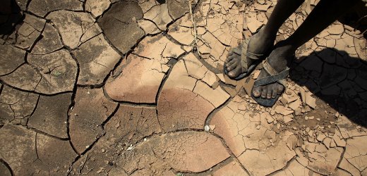 A top-down view of two bare feet standing on a cracked, dry earth surface, indicative of drought conditions. The person is wearing sandals.