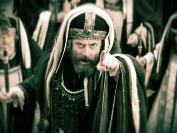 A man dressed in an ornate, ancient robe and headgear, resembling a high priest, is pointing authoritatively towards the camera. His expression is intense and commanding. The image is tinted with a sepia tone, adding a historical atmosphere.