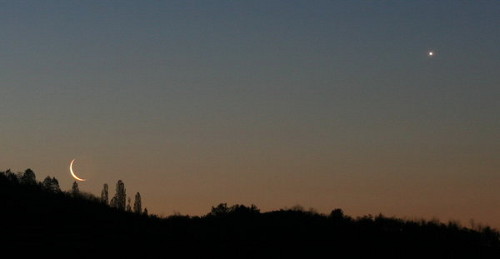 A serene twilight landscape showing a thin crescent moon and a bright celestial body in the sky, hovering above a darkened hill with silhouetted trees.