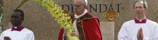 Three clergymen in ceremonial attire, with the central figure in a red robe holding elaborately woven palm fronds, stand before a stone backdrop with engraved lettering, amidst a religious ceremony.