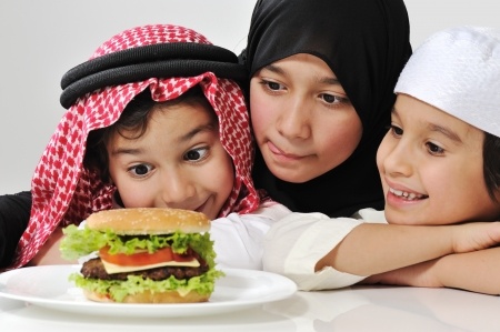 Three young children with Middle Eastern attire, enthusiastically looking at a hamburger on a plate in front of them.