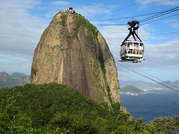 A cable car travels near the summit of a large, steep rock formation surrounded by lush greenery and overlooking a vast ocean.