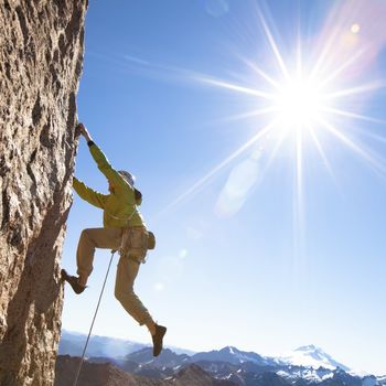 A climber in yellow apparel ascends a steep rock face under a radiant sun near the summit of a mountain range.