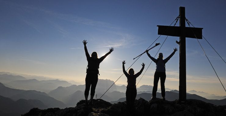 Three hikers celebrate reaching a mountaintop at sunrise, their silhouettes visible against a cross and the softly lit sky, with layers of mountains in the distance.