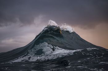 A majestic mountain stands under a dramatic cloudy sky at dusk, its peaks lightly dusted with snow against a fading light backdrop.