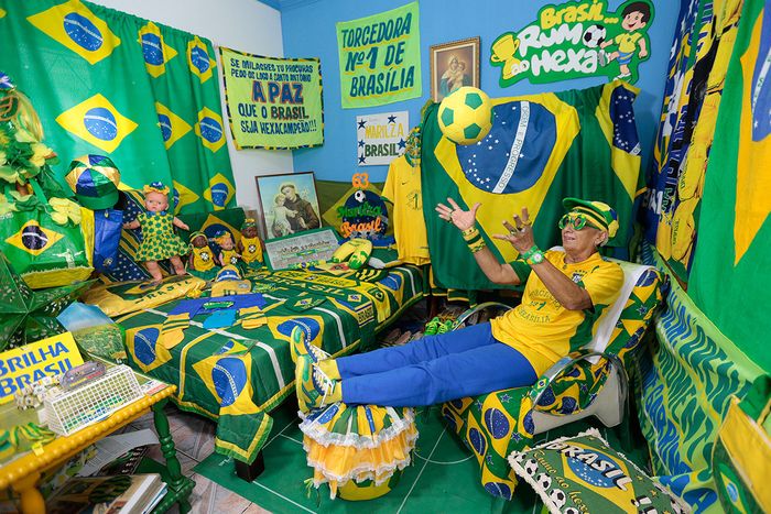 Enthusiastic fan dressed in yellow and green sits in an intricately decorated room filled with various banners, clothing, and memorabilia all themed with the colors of Brazil's national flag. The room exudes a festive, vibrant atmosphere showcasing passion and support for Brazil's national team.