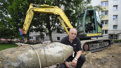 A man in casual clothing is seated on the ground beside a large, unearthed metal object, likely an old bomb, with a yellow excavator parked behind him in an urban area.