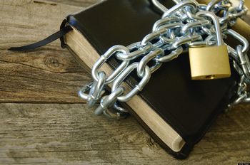 A black book with a gold-trimmed page edge is wrapped with a heavy, silver-chain link and secured with a gold padlock, placed on a rustic wooden surface.