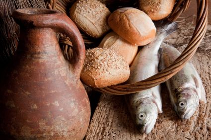 An ancient clay pitcher stands beside a basket filled with various fresh bread loaves and two whole fish, all resting on a rustic wooden surface.