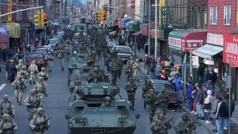 A battalion of soldiers in camouflage uniforms marches down a city street, accompanied by several armored military vehicles. Civilians watch from the sidewalks lined with diverse shops.