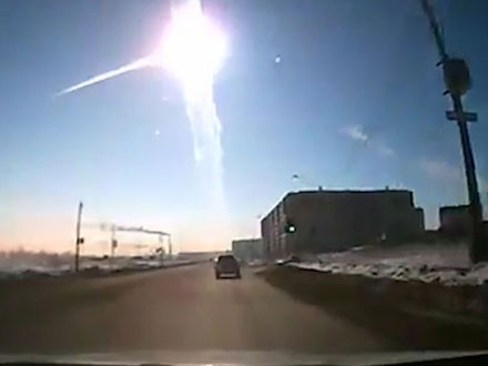A meteor streaks vividly across a clear blue sky, illuminating the scene with a brilliant trail, over a snowy roadside landscape featuring buildings and moving vehicles.