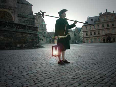 A person dressed in historical attire stands in a cobblestone square at dusk, holding a lantern that glows warmly and a long pole. The surrounding architecture includes classical buildings and an overcast sky suggesting an early modern European city.