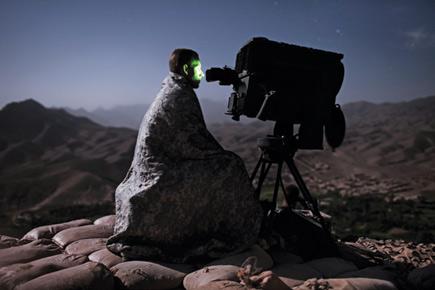 An individual is seated outdoors at night, enveloped in a rock-pattern cloak, looking at the heavens through a large telescope positioned on a rugged terrain with distant mountains under a starlit sky.