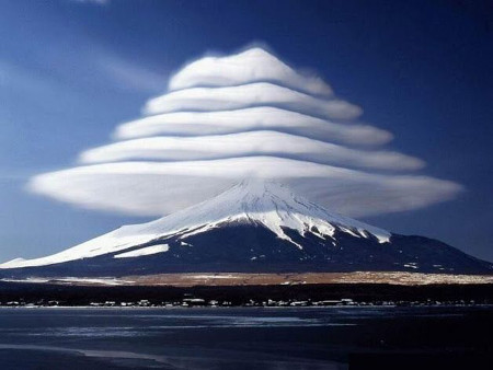 A snow-capped mountain with multiple lenticular clouds stacked above it, resembling a celestial phenomenon, contrasting with a clear blue sky and surrounded by a barren landscape.