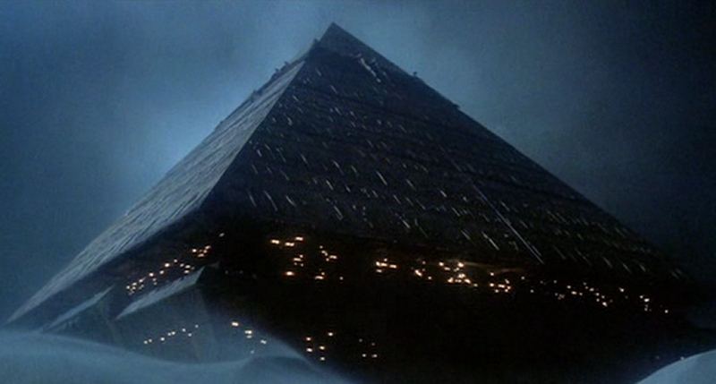 An illuminated, large pyramidal structure emerging from a dark, misty body of water under a night sky, reminiscent of ancient celestial observatories used to study the heavens.