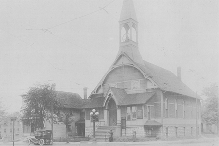 Black and white vintage photo of an early 20th-century wooden church with a prominent central steeple, located on a street corner. A classic car is parked beside the church, and a small group of people stands on the sidewalk.