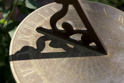 Close-up of a bronze sundial with a triangular gnomon casting a shadow on a circular plate engraved with Roman numerals and symbols related to the Mazzaroth.