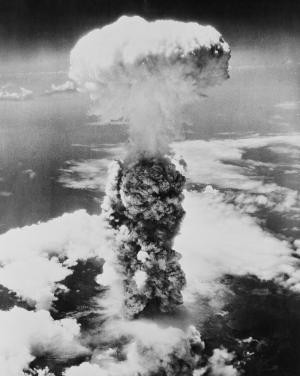 Historic black and white photo of an enormous mushroom cloud from an atomic bomb explosion rising above a dense layer of clouds. The cloud extends high into the sky, creating a stark and dramatic contrast against the surrounding atmosphere.