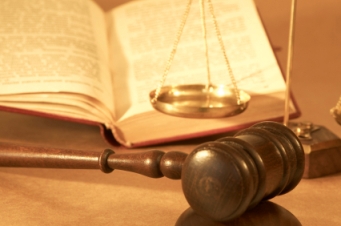 A wooden gavel and a set of brass scales on a desk, symbolizing justice and balance, next to an open book with visible text.