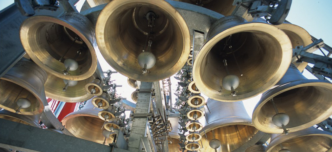 An array of large bronze bells suspended in a steel framework, viewed from below. Each bell has a visible clapper and varying diameters, reflecting light differently.