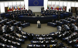 A wide-angle view of a large, semi-circular assembly hall where numerous delegates are seated at desks arranged in a curved pattern. A central speaker stands at the podium, addressing the assembly under a large circular emblem with gold stars on a blue background.