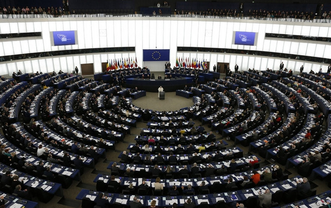 A wide view of a large, hemicycle parliamentary chamber during a session, with numerous delegates seated in a semi-circular arrangement. A central podium and flags of various nations along with the EU flag are visible in the background.