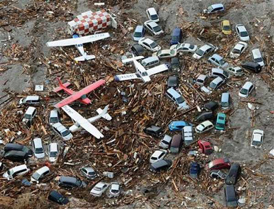An aerial view showing a collection of toy vehicles including cars and planes scattered on a surface covered with debris and small sticks.