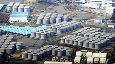 Aerial view of an industrial site with numerous large cylindrical storage tanks symmetrically arranged, surrounded by various buildings, vehicles, and construction activity.