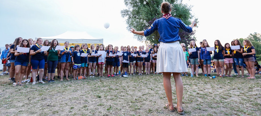 A group of excited female scouts holding papers listens to a leader directing them outdoors, beneath serene skies, on a grassy area.
