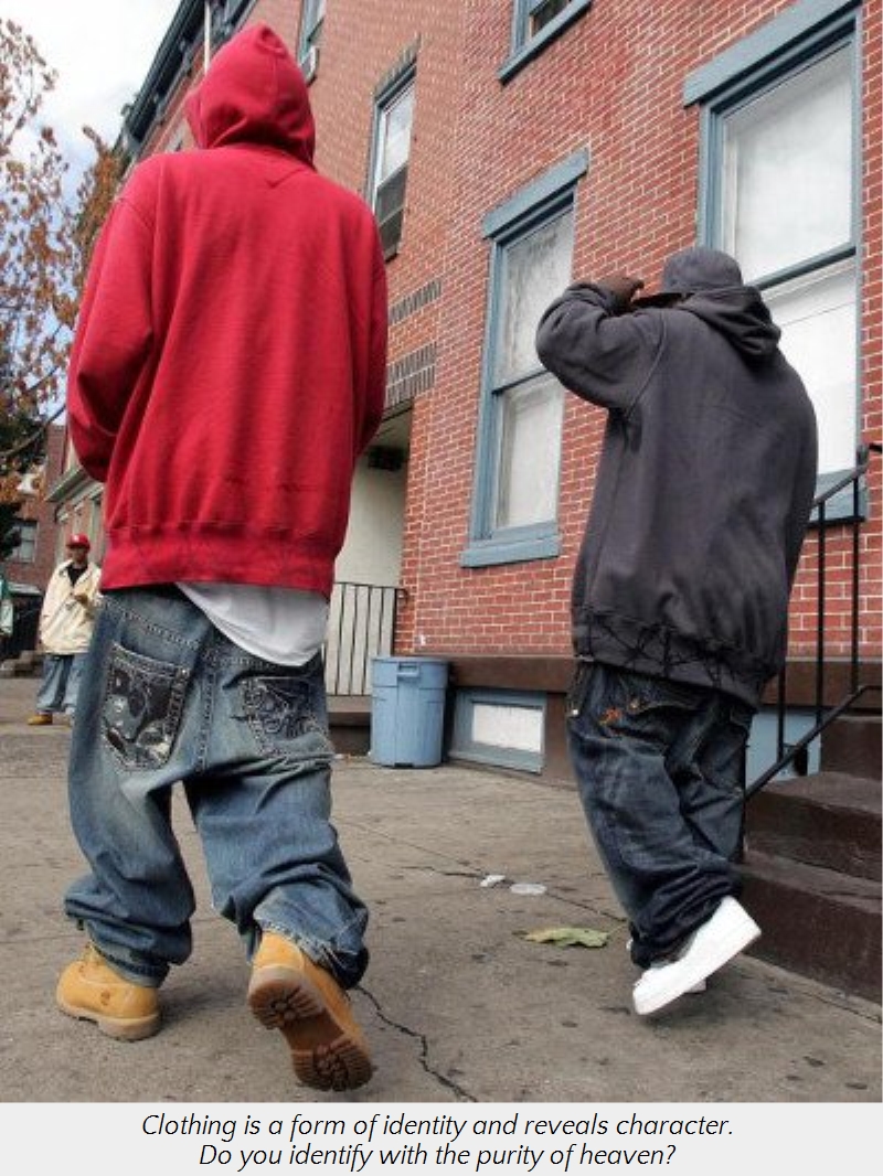 Two young men walking on a city street. The man on the left wears a red hooded sweatshirt, and the man on the right is in a black hoodie. Both wear baggy jeans and sneakers. They are passing by red-brick buildings with windows.