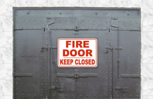 A large gray metal door with riveted panels and a red and white sign that reads "FIRE DOOR KEEP CLOSED" mounted on it, against a light gray wall.