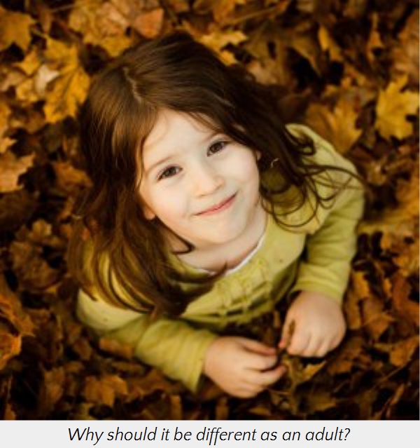 A young child with a joyful expression lies in a bed of autumn leaves, gazing upward. The child's hair is long and brown, and they wear a light green shirt. The leaves are various shades of gold and brown.