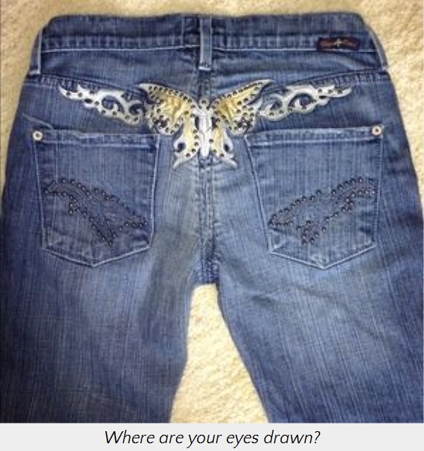A pair of denim jeans laid out flat, featuring an intricate celestial butterfly design in white and gold at the waistband, with sparkling embellishments on the back pockets.
