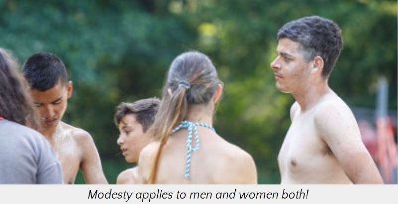 Group of young adults standing outdoors in a park, engaged in a casual conversation. Two shirtless men and one woman with her back turned, seen from the shoulders up, are visible. Green trees blur softly in the background.
