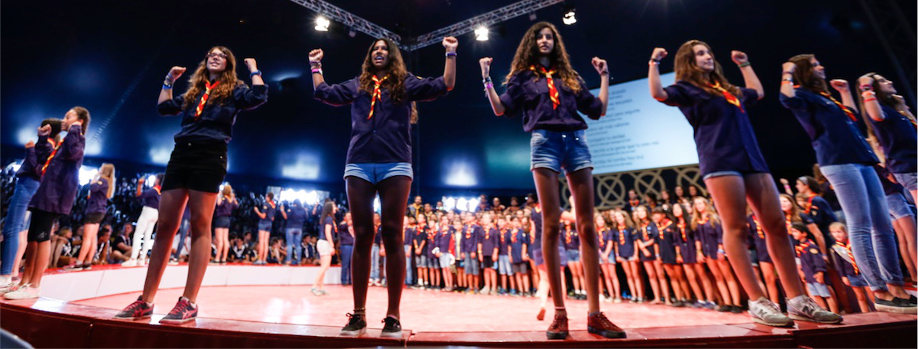 A group of enthusiastic students raising their hands in jubilation, standing on a circular stage at a gathering, with many peers supporting them in the background, all in a large auditorium under artificial lighting.