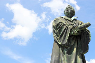 A bronze statue of a bearded man in robes standing under a clear blue sky, with his arms crossed and looking upwards.