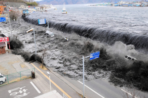 Aerial view of a massive tidal wave crashing over a sea wall onto a coastal road, with buildings and parked vehicles near the shore.