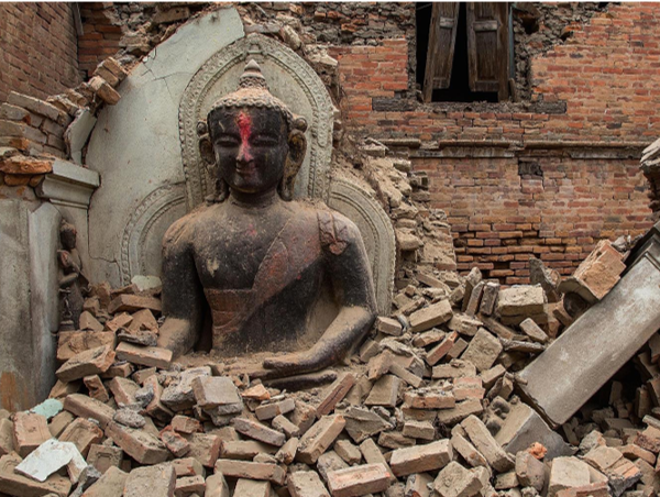A serene stone sculpture of a seated figure surrounded by a halo-like arch, rises amidst a pile of brick rubble. Evidently enduring yet partly shielded by the arch, the figure carries signs of age and weathering, hinting at historical and cultural significance lost in an urban setting where the brick architecture suffers destruction.