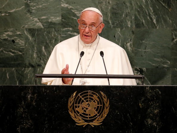 A man in white religious attire speaks from a podium embellished with the emblem of the United Nations, set against a large green marble background.